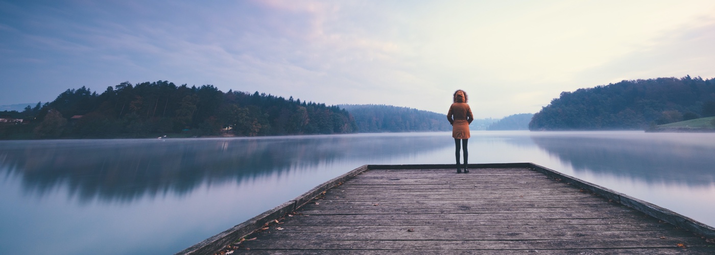 Back view of young woman with hoodie and hand in pockets standing on dock in front of river having double exposure